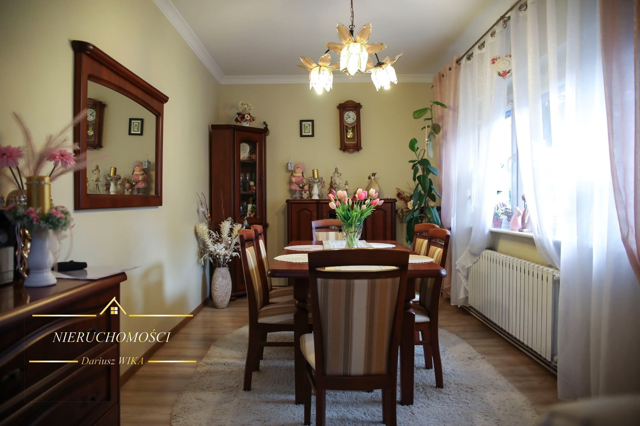 Jasna jadalnia z drewnianym stołem i krzesłami, bukietem tulipanów, ozdobami i oknem z firanami. Ciepłe oświetlenie i przytulna atmosfera.