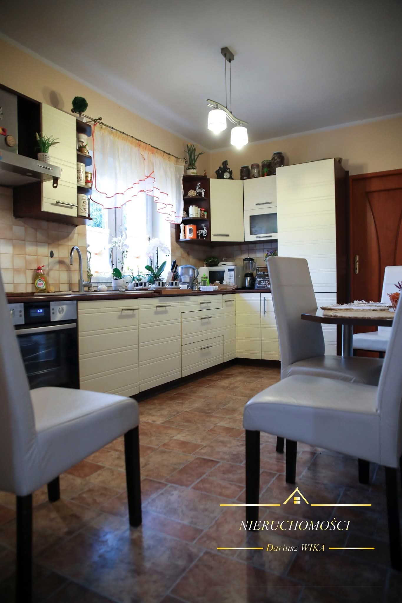 Jasna kuchnia z beżowymi frontami, brązowym blatem i płytkami podłogowymi. Widoczne krzesła i stół. Okno z firanką w tle. Oświetlenie sufitowe.