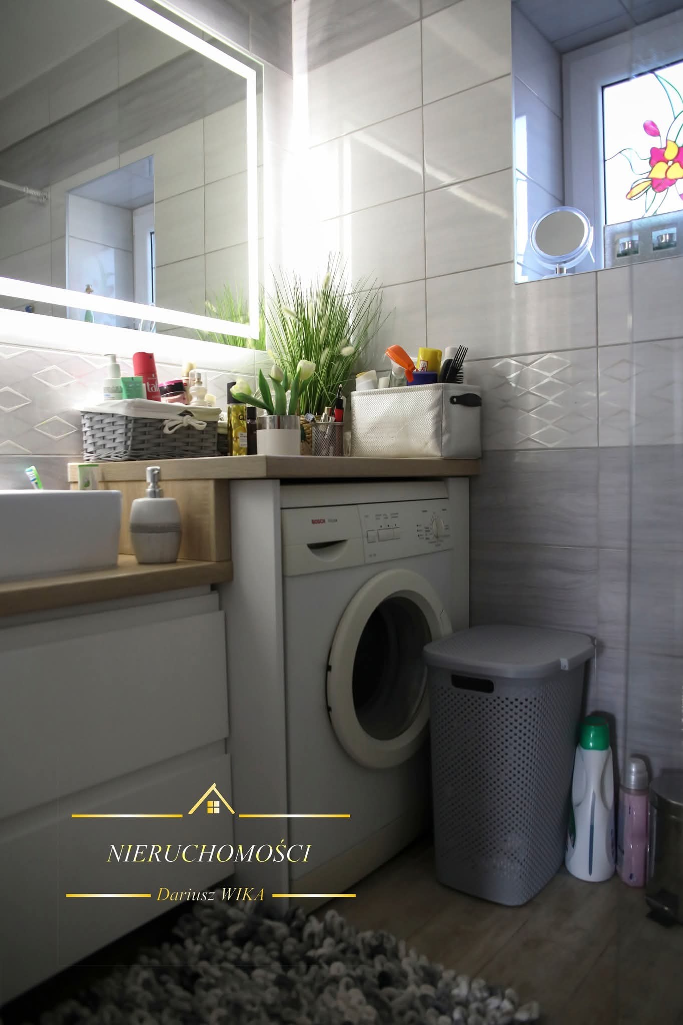Jasna łazienka z pralką Bosch, umywalką i lustrem z podświetleniem LED. Na blacie dekoracje, a na ścianie płytki w odcieniach szarości. W tle okno z witrażem.