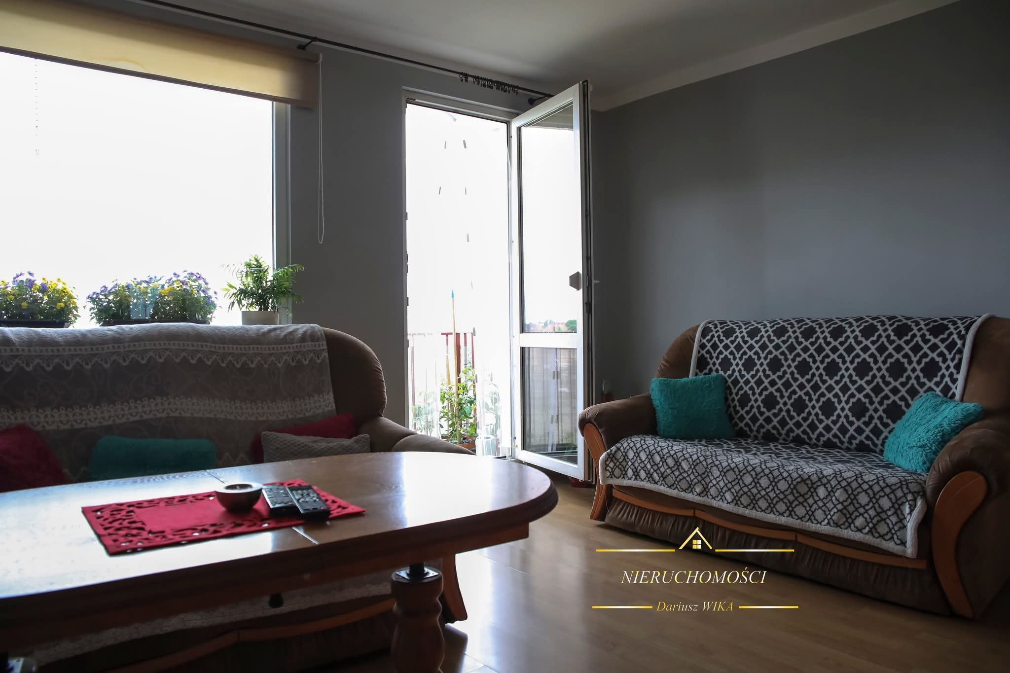 Jasne wnętrze salonu z balkonem, dwa brązowe fotele z poduszkami, drewniany stolik kawowy z pilotami, kwiaty w oknie, parkiet. Widok z mieszkania na zewnątrz.