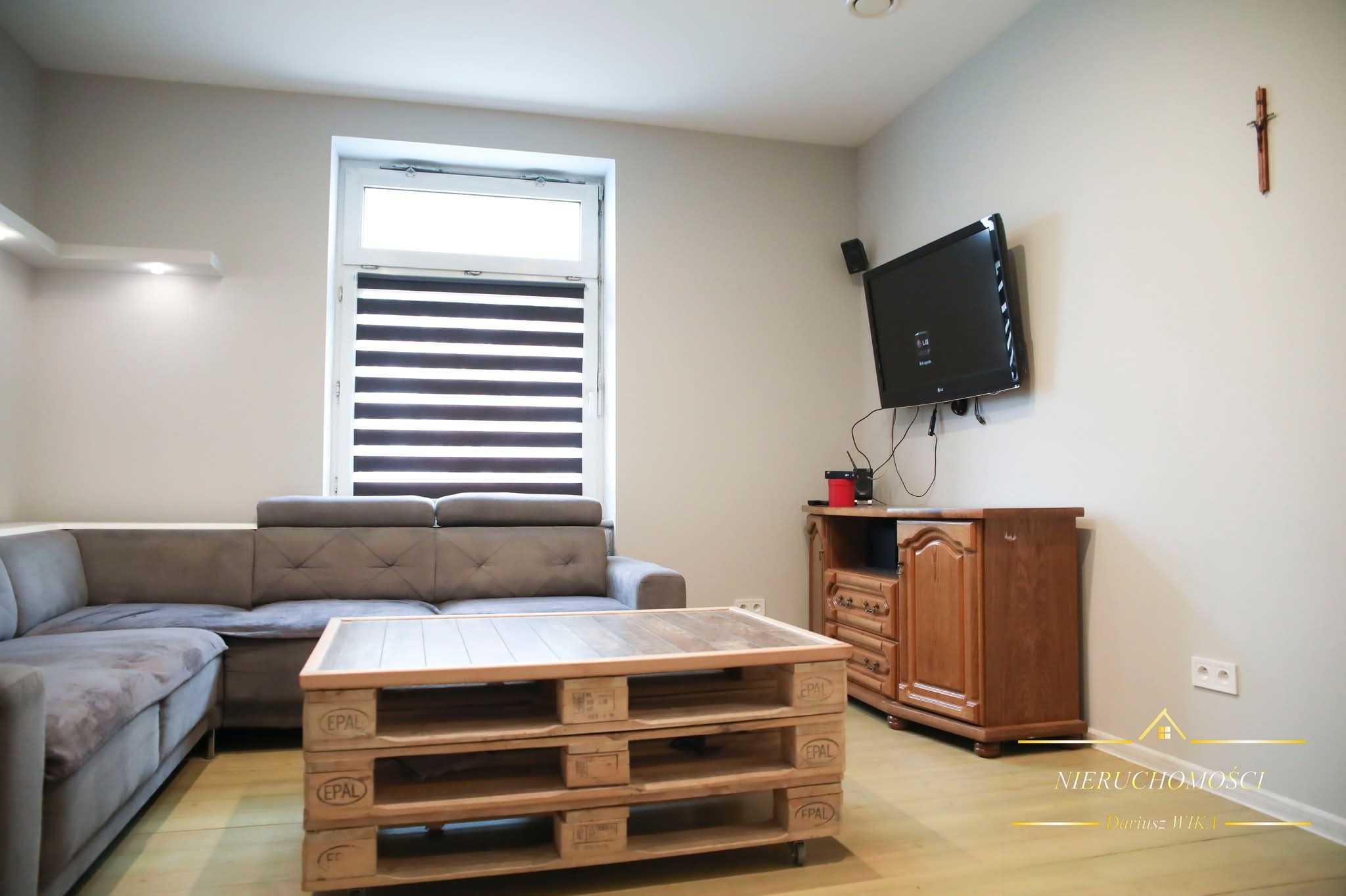 Jasne wnętrze salonu w Międzyrzeczu z szarą sofą narożną, stolikiem z palet i drewnianą komodą pod telewizorem. Okno z roletami w tle.