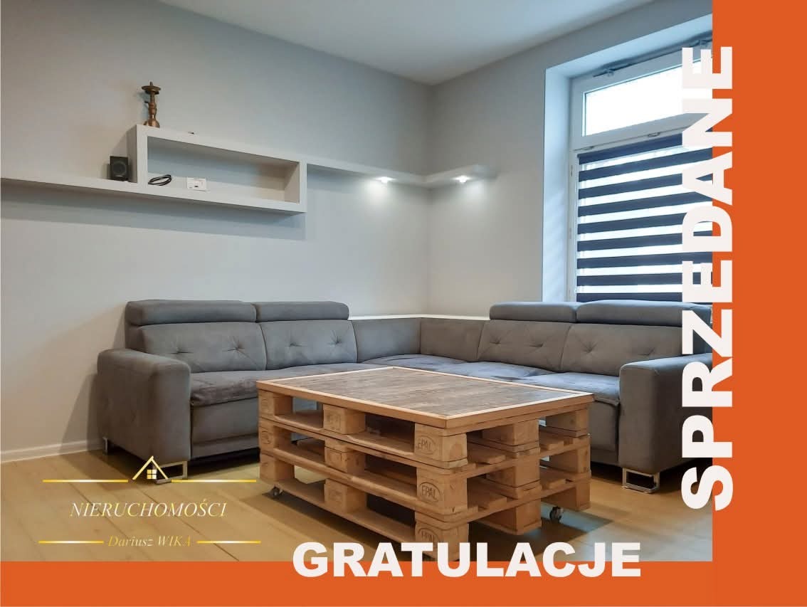 Jasne wnętrze salonu z szarą sofą narożną i stolikiem z palet. Na ścianie półki, okno z żaluzjami. Po prawej stronie napis 'Sprzedane' i 'Gratulacje'.
