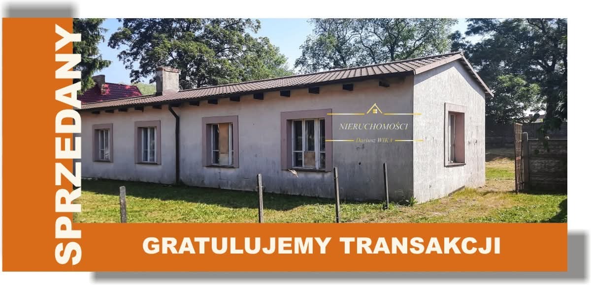 Dom parterowy z ogrodem, otynkowany na biało, z brązowym dachem. Na zdjęciu widnieje pomarańczowy baner z napisem 'Sprzedany' i 'Gratulujemy transakcji'.