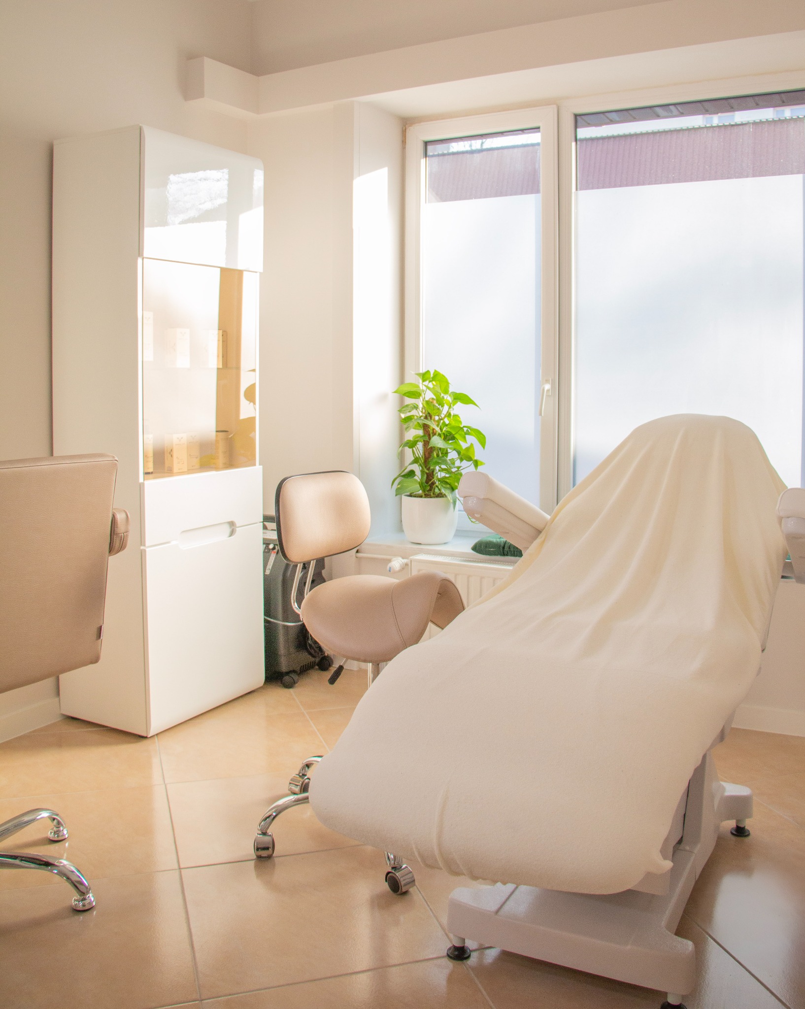 Jasne wnętrze gabinetu kosmetycznego z fotelem zabiegowym przykrytym kremowym pokrowcem, obok krzesło na kółkach i biała szafka ekspozycyjna oraz roślina doniczkowa na parapecie.