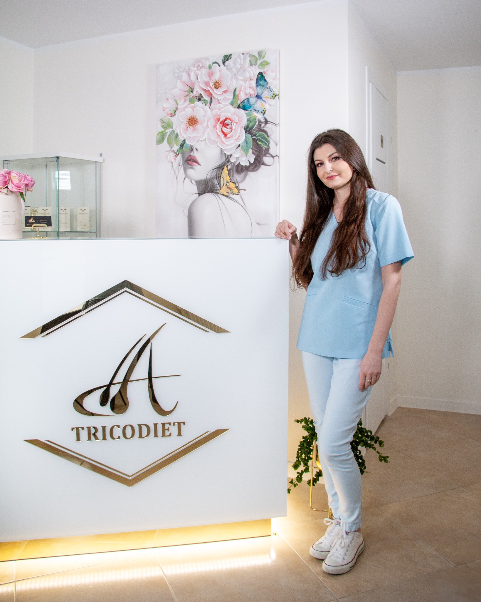 Recepcja gabinetu z logo Tricodiet i portretem kobiety z kwiatami, pracownik w błękitnym uniformie opiera się o ladę