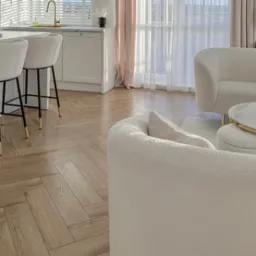 Jasna, drewniana podłoga ułożona w jodełkę francuską w eleganckim salonie z białymi meblami i złotymi akcentami.