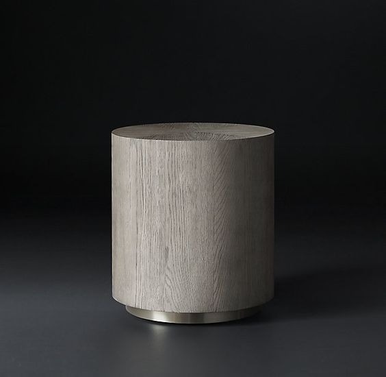 Cylindryczny stolik nocny z jasnego drewna z widocznym usłojeniem, osadzony na metalowej podstawie w kolorze szczotkowanego niklu, na ciemnym tle.