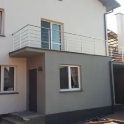 Nowoczesna balustrada ze stali nierdzewnej na balkonie domu jednorodzinnego z jasną elewacją, widoczne puszki po farbie u podstawy schodów.