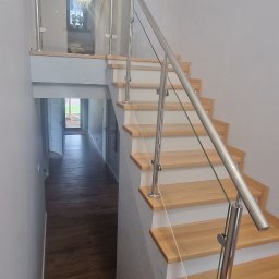 Mariusz Dziedzic Lux-Inox - Klatka schodowa z drewnianymi stopniami i balustradą ze stali nierdzewnej z wypełnieniem ze szkła, widok z góry na dół, w tle widoczny korytarz z ciemną podłogą i białymi drzwiami.