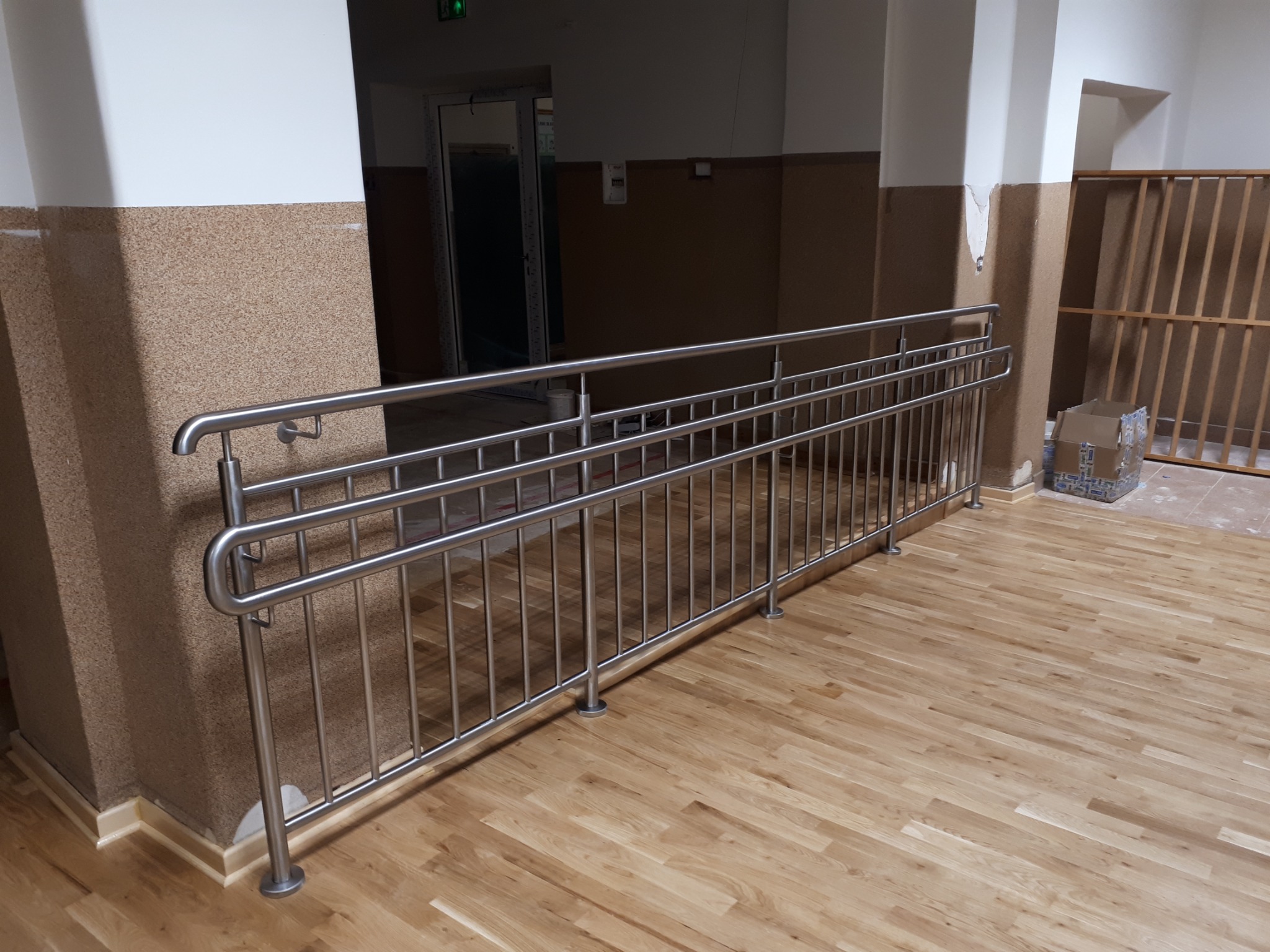 Nierdzewna balustrada schodowa z pionowymi prętami, zamontowana wewnątrz budynku na drewnianej podłodze, z widocznym fragmentem korytarza i ścianą wykończoną w dwóch odcieniach brązu.