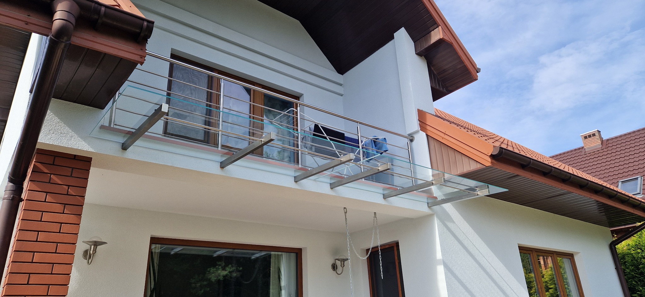 Balkon z balustradą ze stali nierdzewnej i szkła, zadaszony szkłem, widok z dołu na elewację domu z oknami i brązowym dachem.