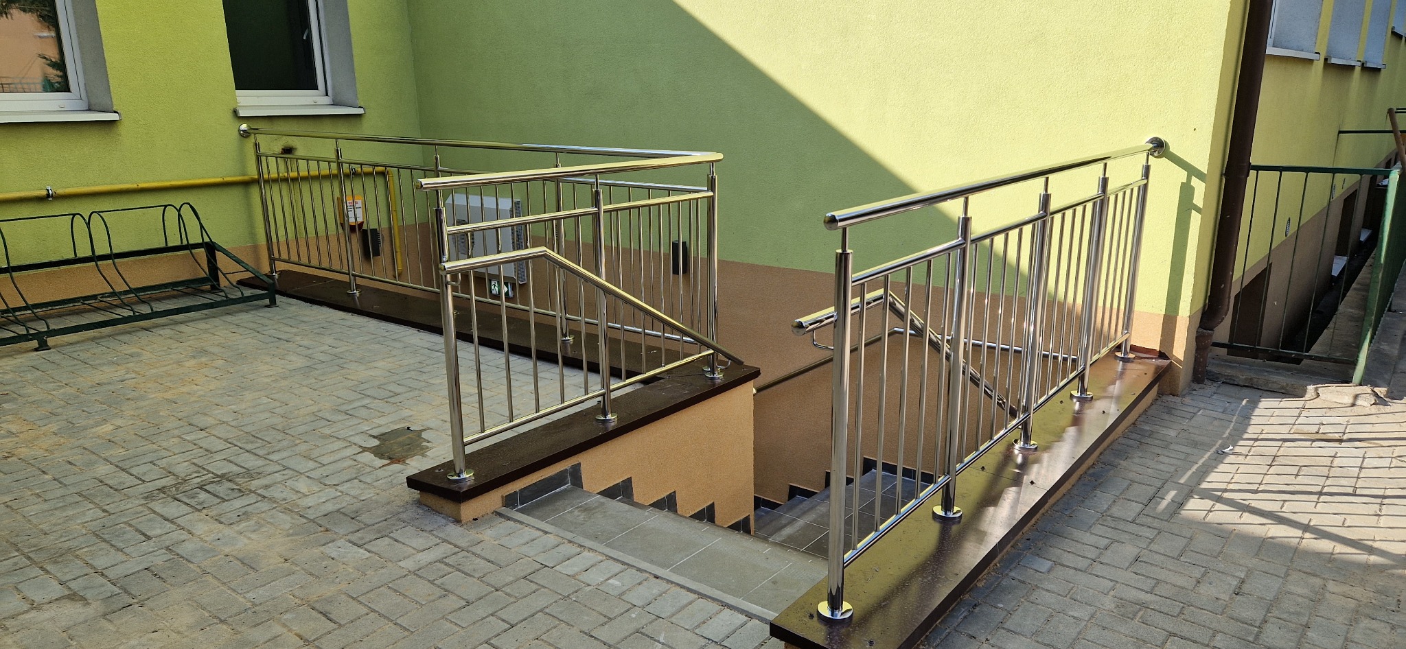 Zewnętrzne schody do piwnicy z metalowymi poręczami ze stali nierdzewnej, widok z góry na kostkę brukową i zieloną elewację budynku.