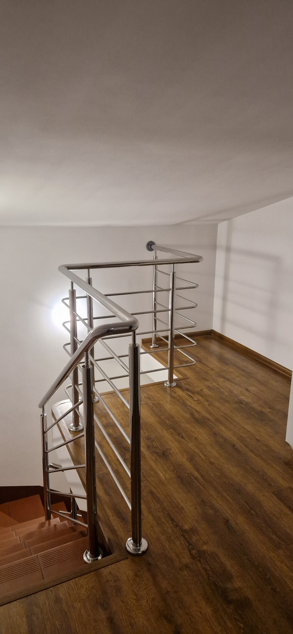 Wnętrze domu z drewnopodobną podłogą i schodami wykończonymi terakotą, zabezpieczonymi nowoczesną, chromowaną balustradą z poziomymi prętami.