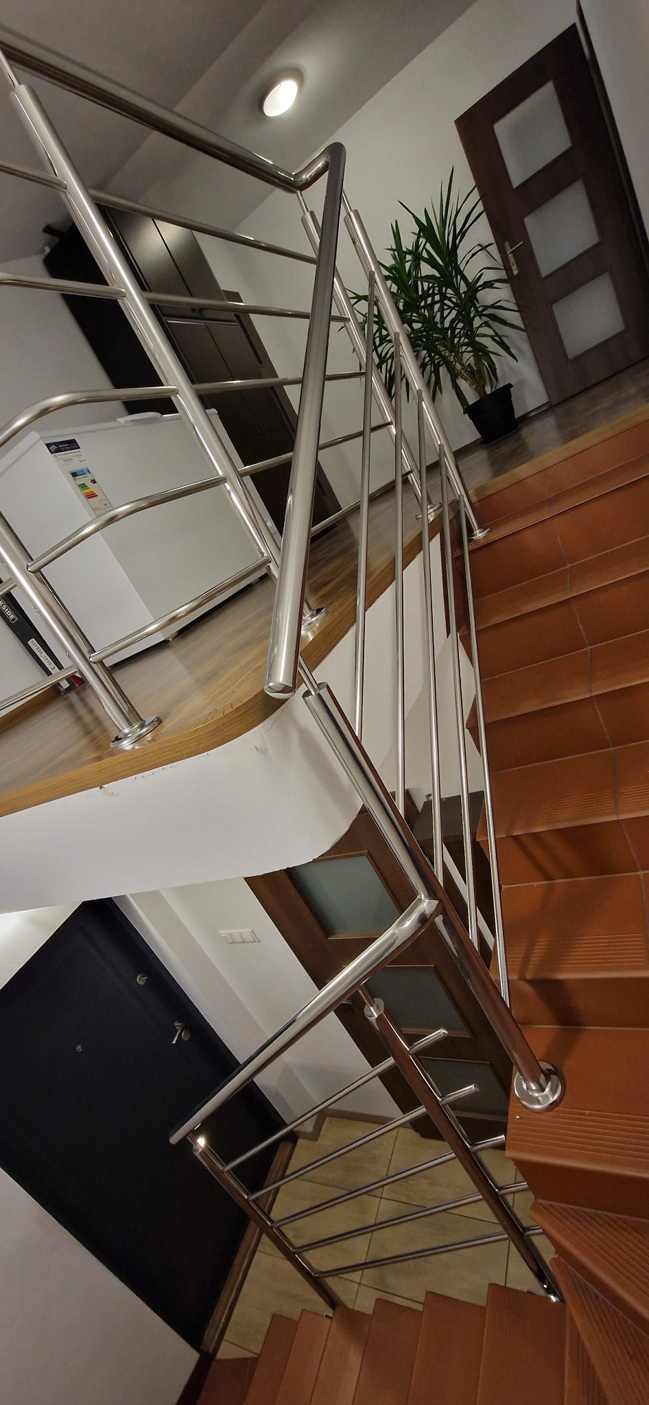 Wnętrze domu z drewnianymi schodami z brązowymi płytkami, zabezpieczonymi nowoczesną balustradą ze stali nierdzewnej o minimalistycznym designie, widok z dołu na górę, z fragmentem rośliny...