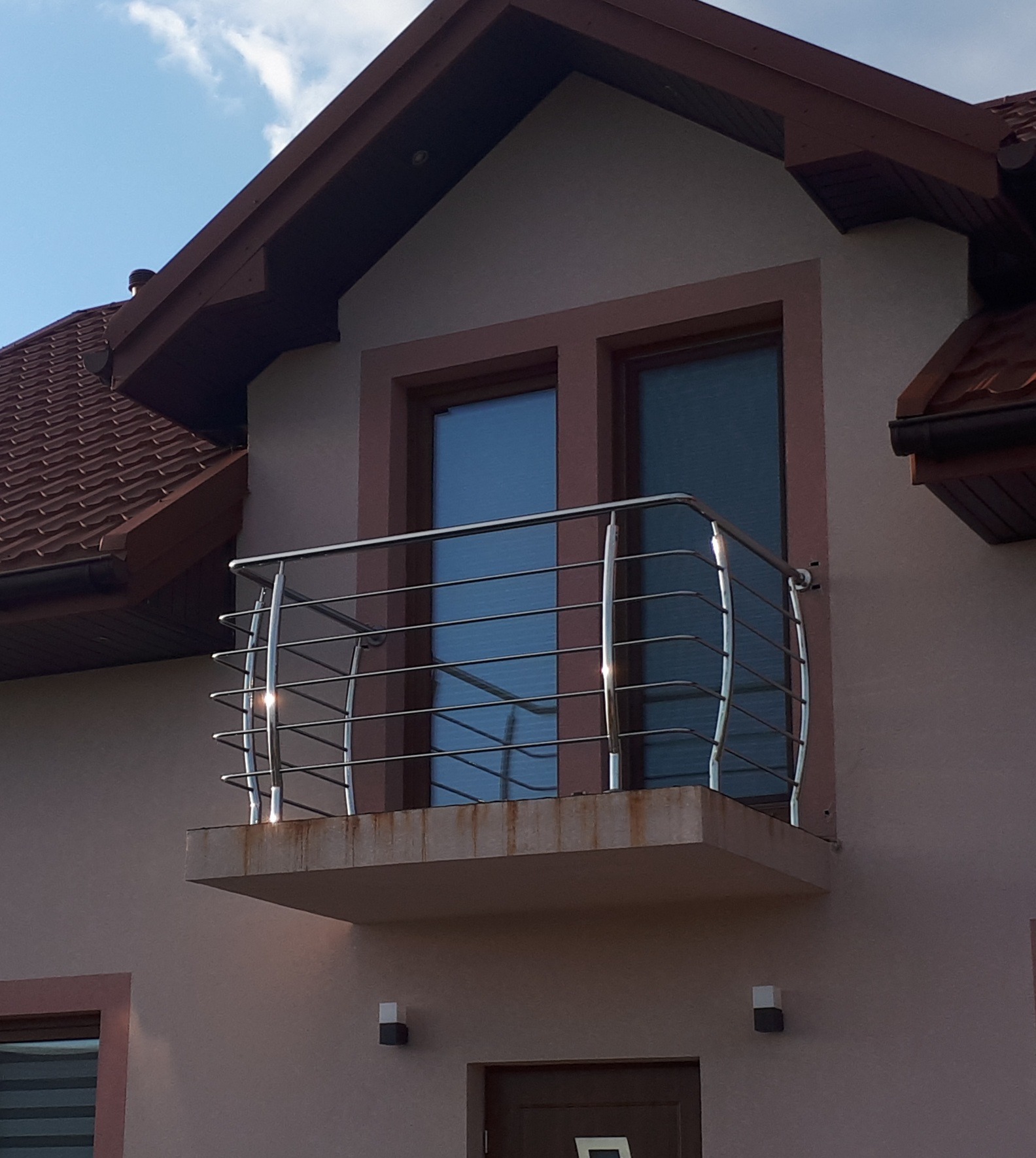 Balkon z balustradą ze stali nierdzewnej na tle dwuspadowego dachu i błękitnego nieba.