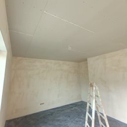 Jacek Staniszewski Usługi Ogólnobudowlane - Pokój w trakcie prac wykończeniowych: ściany wstępnie przygotowane, sufit z płyt kartonowo-gipsowych, zabezpieczona podłoga i drewniana drabina. Widoczne ślady po szpachlowaniu.