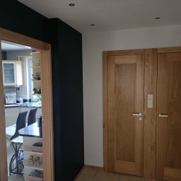 Jacek Staniszewski Usługi Ogólnobudowlane - Nowoczesne wnętrze z czarną ścianą i drewnianą stolarką drzwiową, widok na kuchnię z białymi meblami przez drewnianą ościeżnicę.