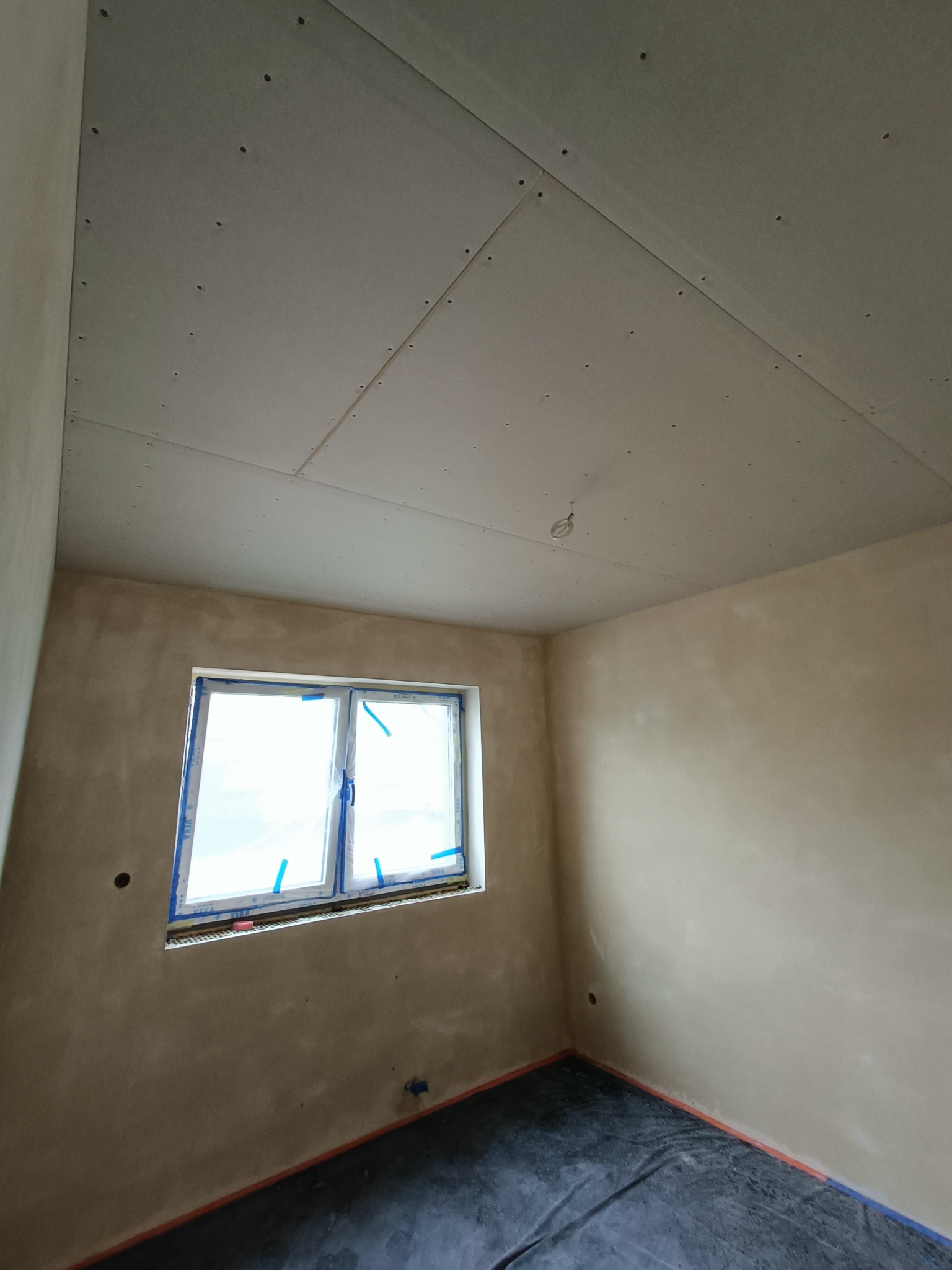 Pokój w trakcie prac wykończeniowych: ściany po nałożeniu warstwy szpachli, sufit z zamontowaną płytą karton-gipsem, okno zabezpieczone folią, podłoga przykryta folią ochronną.