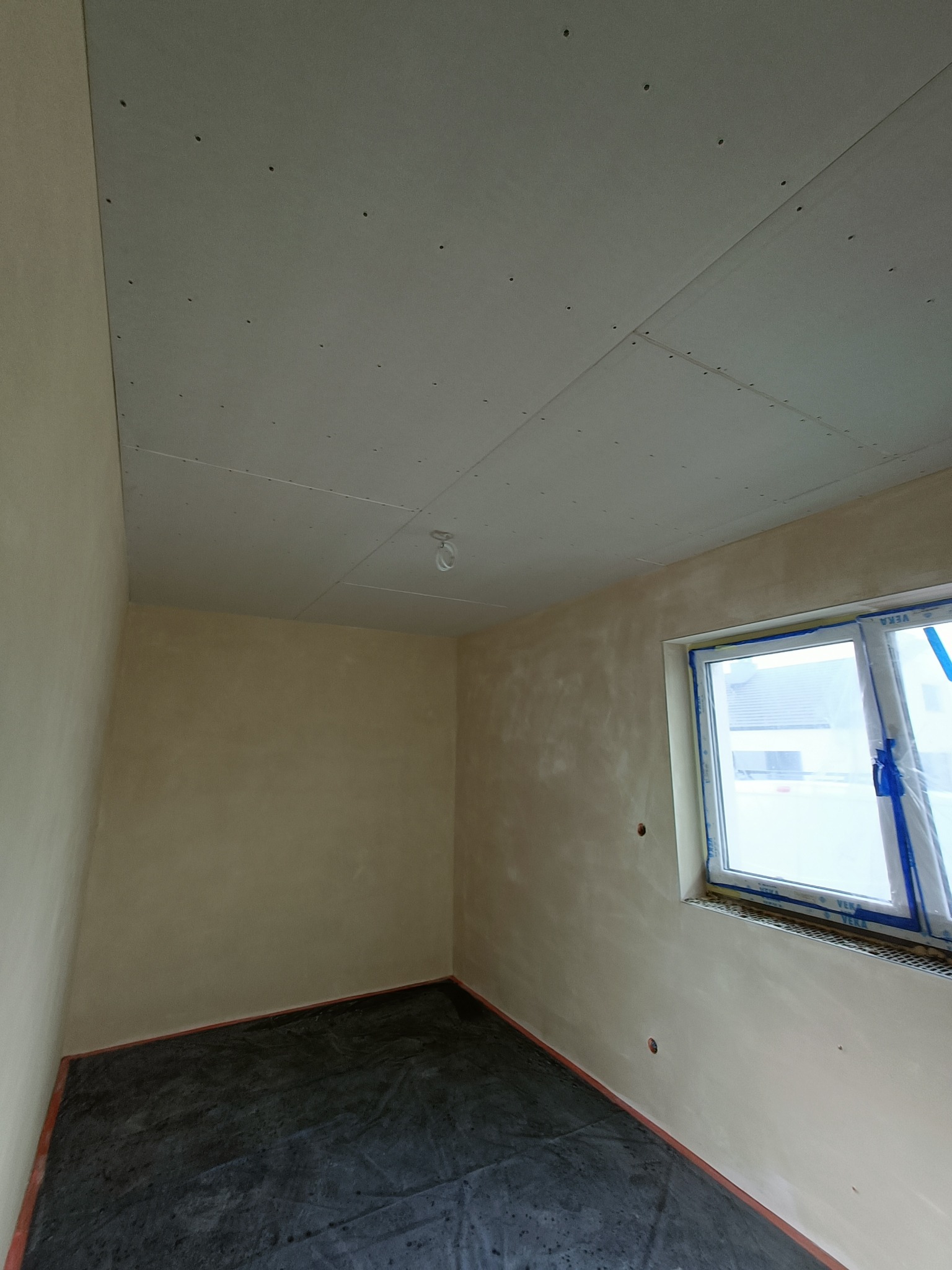 Surowe wnętrze w trakcie prac wykończeniowych: ściany po wstępnym pokryciu, sufit z zamontowaną płytą gipsowo-kartonową z widocznymi otworami po wkrętach, okno zabezpieczone folią ochronną.