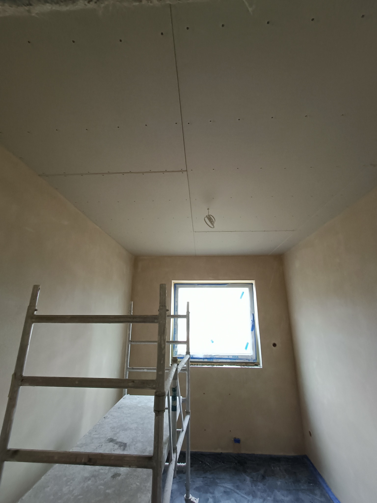 Wnętrze w trakcie prac: sufit z płyt kartonowo-gipsowych z widocznymi łączeniami, ściany przygotowane pod malowanie, okno zabezpieczone folią, metalowe rusztowanie w pomieszczeniu.