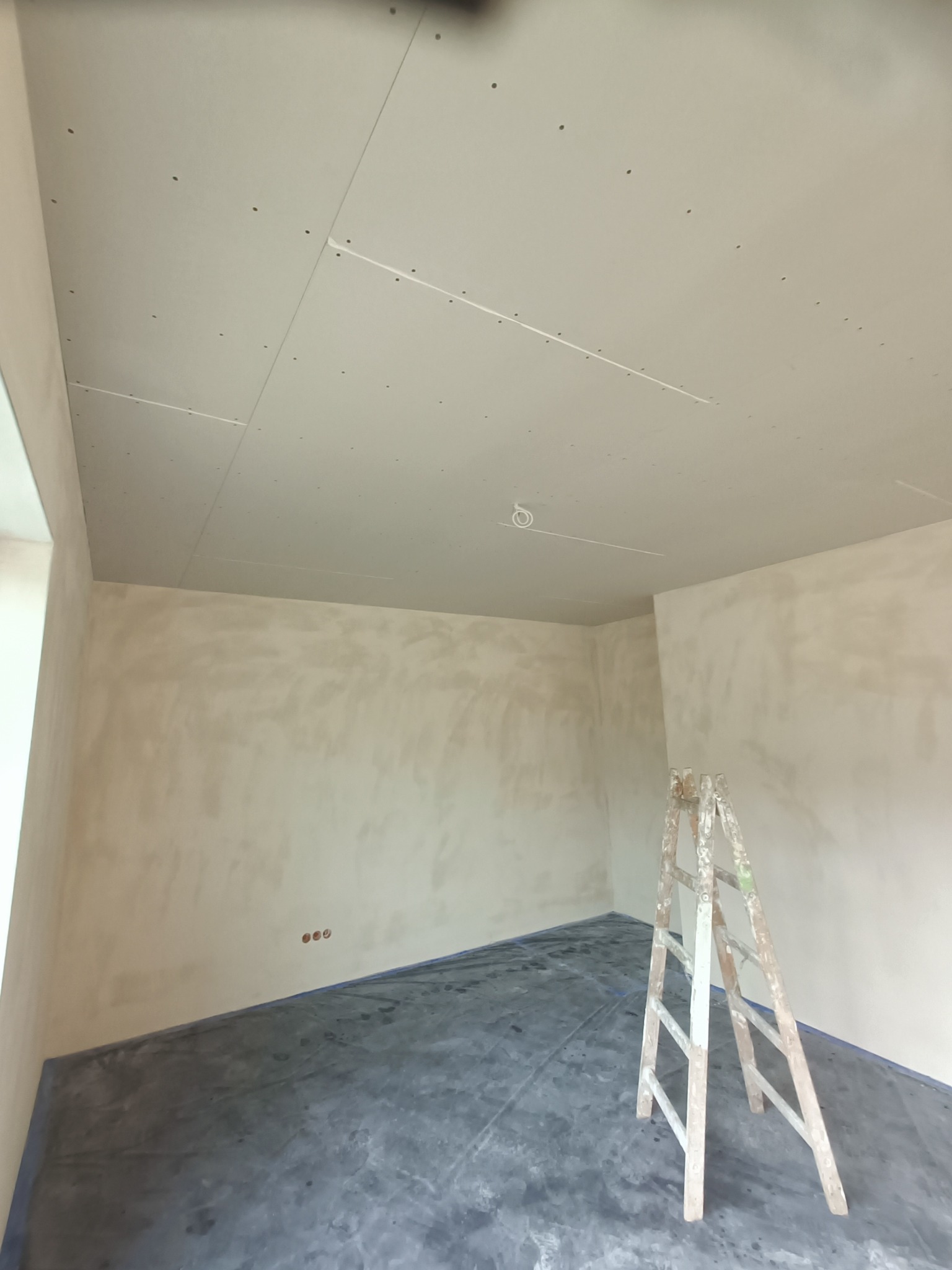 Pokój w trakcie prac wykończeniowych: ściany wstępnie przygotowane, sufit z płyt kartonowo-gipsowych, zabezpieczona podłoga i drewniana drabina. Widoczne ślady po szpachlowaniu.
