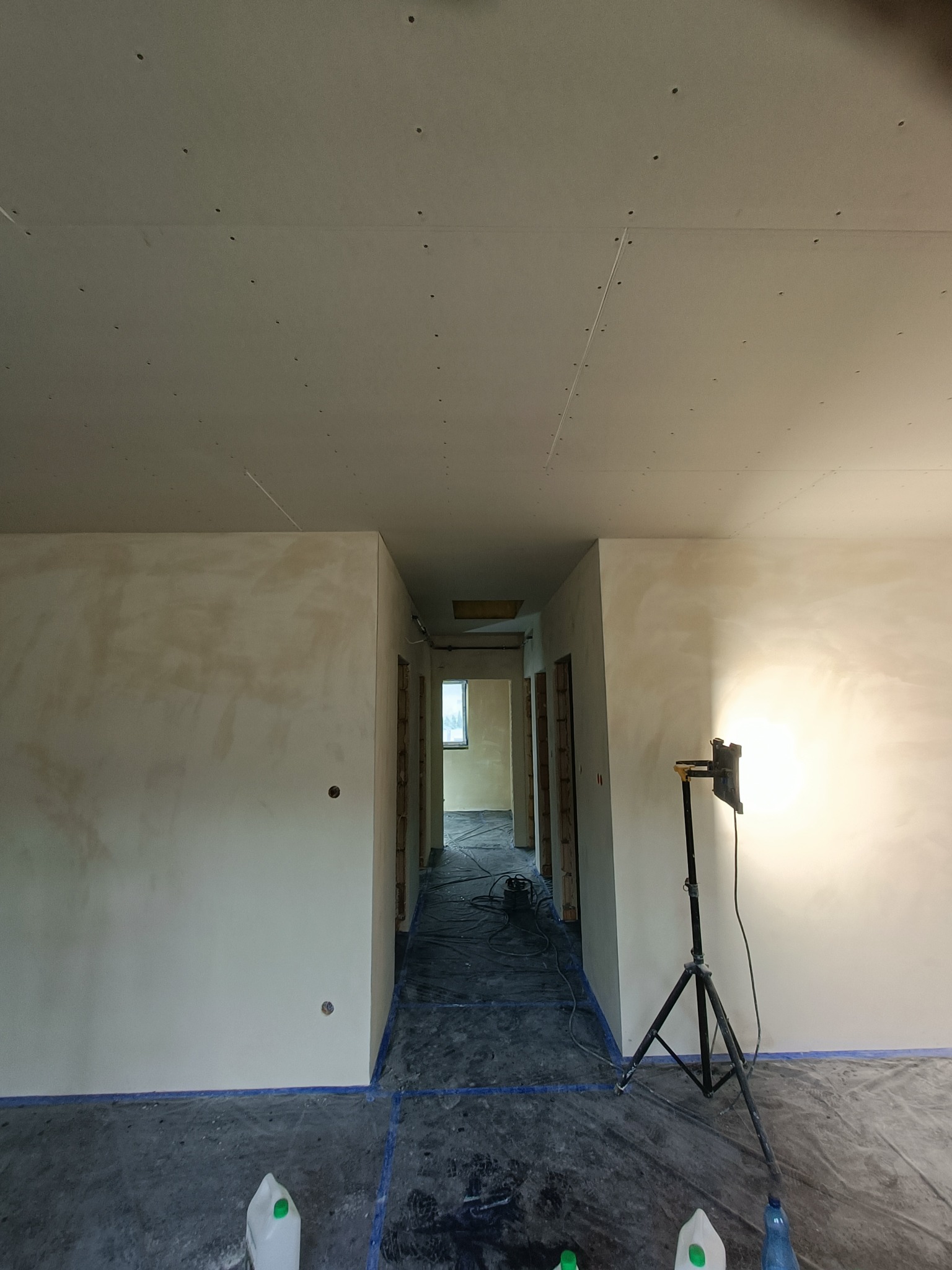 Korytarz w trakcie prac wykończeniowych: ściany po szpachlowaniu, sufit z płyt karton gips, podłoga zabezpieczona folią, widoczny reflektor na statywie i butelki z płynami.