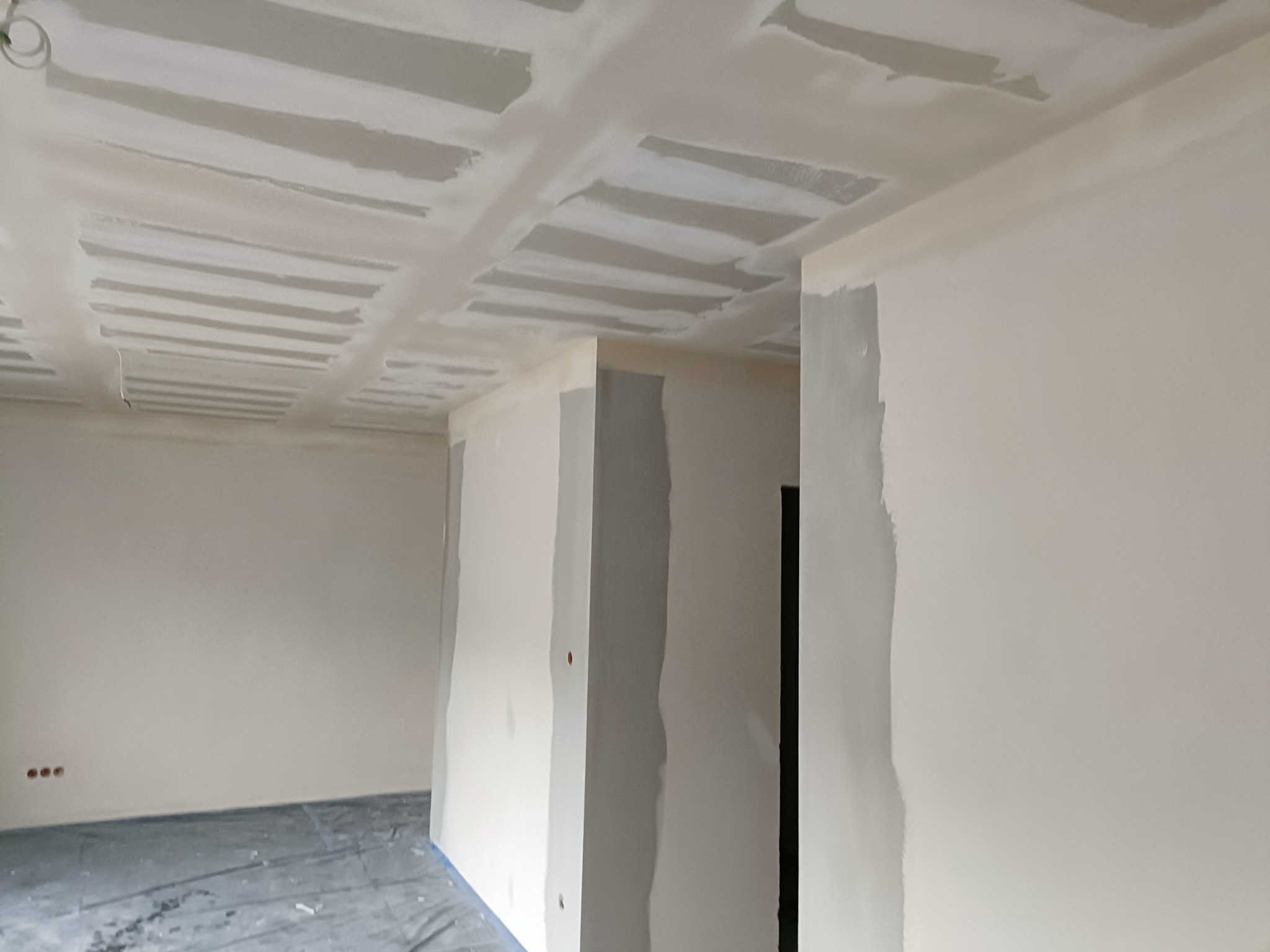 Wnętrze w trakcie prac wykończeniowych: sufit z widoczną konstrukcją i nałożoną warstwą masy szpachlowej, ściany częściowo pokryte masą, podłoga zabezpieczona folią.