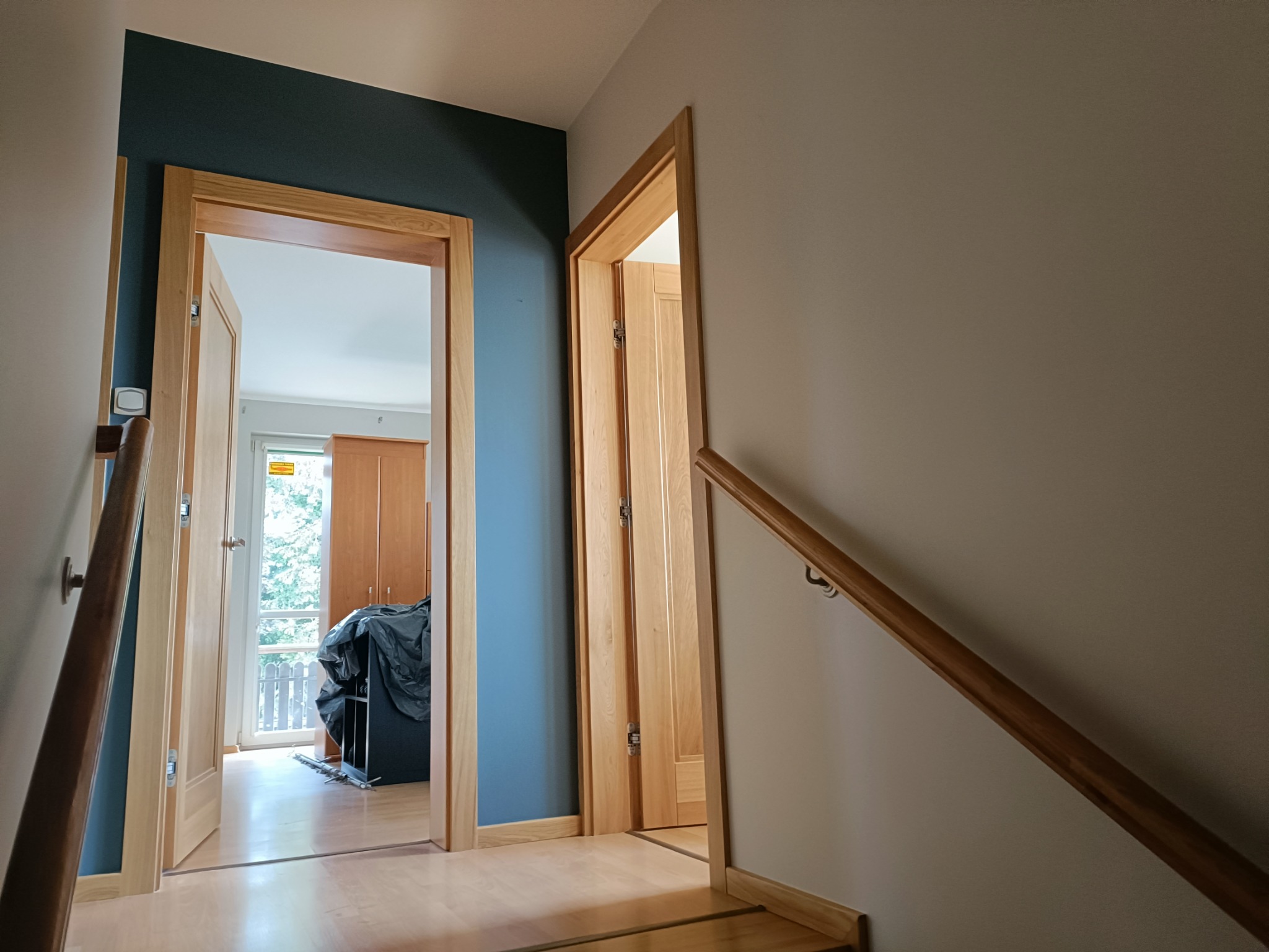 Korytarz po remoncie z dwoma otwartymi drewnianymi drzwiami, ściany pomalowane na szaro i ciemny niebieski, widoczna drewniana balustrada schodów.