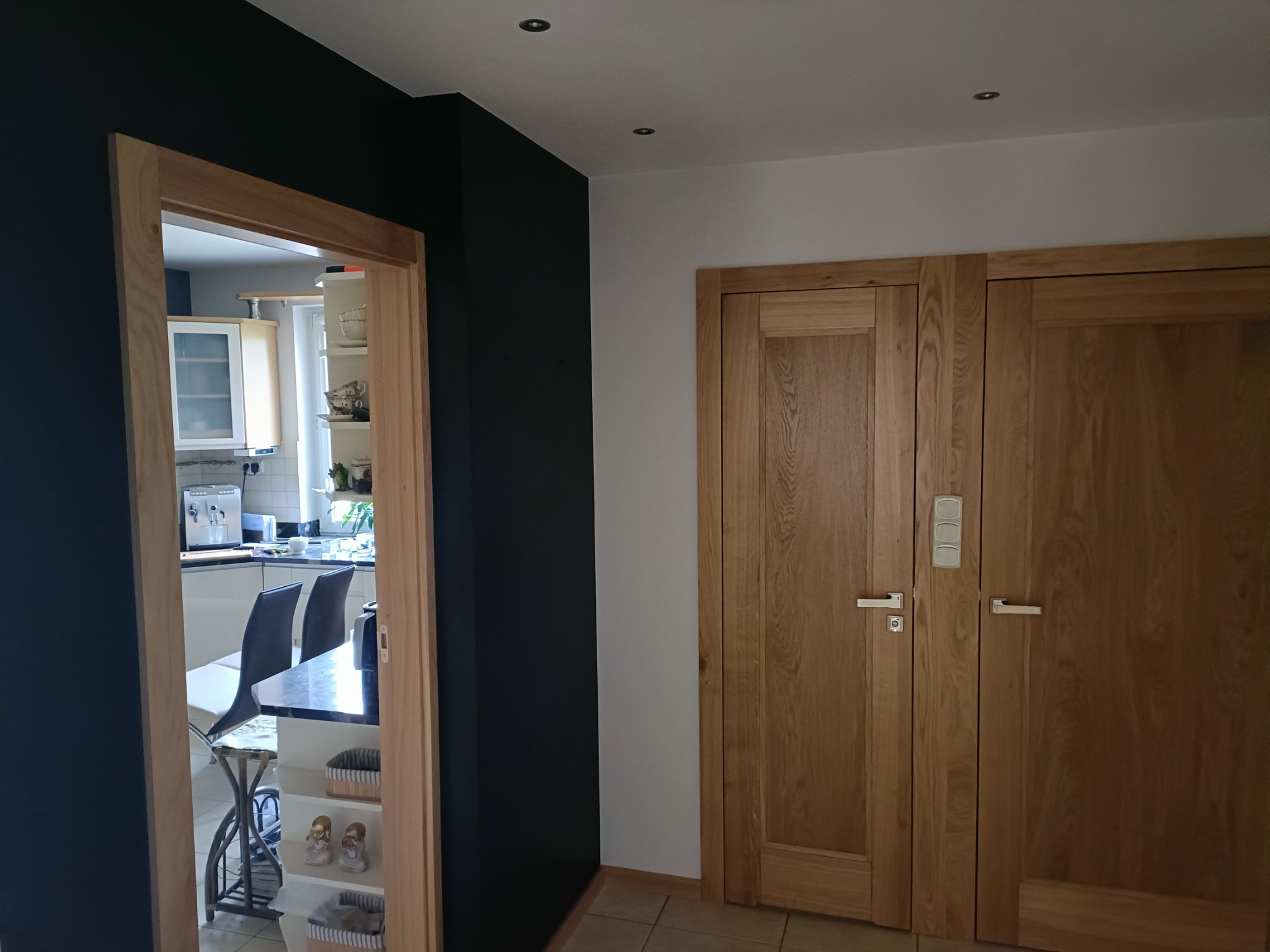 Nowoczesne wnętrze z czarną ścianą i drewnianą stolarką drzwiową, widok na kuchnię z białymi meblami przez drewnianą ościeżnicę.