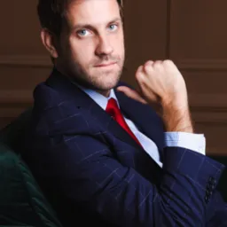 Portret mężczyzny w granatowym garniturze w kratę i czerwonym krawacie, opierającego się o zieloną sofę, z poważnym wyrazem twarzy.