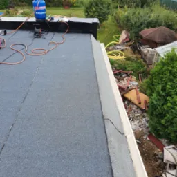 Nowo pokryty, płaski dach z papy termozgrzewalnej, widoczny niebieski zbiornik gazowy WASBRUK i narzędzia dekarskie, w tle zielony ogród z elementami demontażu konstrukcji dachowej.