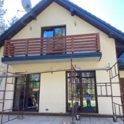 Dom jednorodzinny z beżową elewacją, drewnianym balkonem i ciemną dachówką, częściowo zasłonięty rusztowaniem malarskim. Widoczna antena satelitarna na dachu.