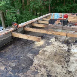 Demontaż starego dachu z widocznymi elementami konstrukcji drewnianej, ceglaną obmurówką, piłą spalinową i butelkami wody mineralnej na tle zielonego lasu.