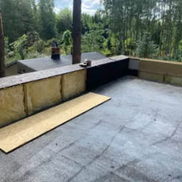 Poddasze w trakcie ocieplania wełną mineralną i obijania płytami OSB, widok na drzewa i dach sąsiedniego budynku.