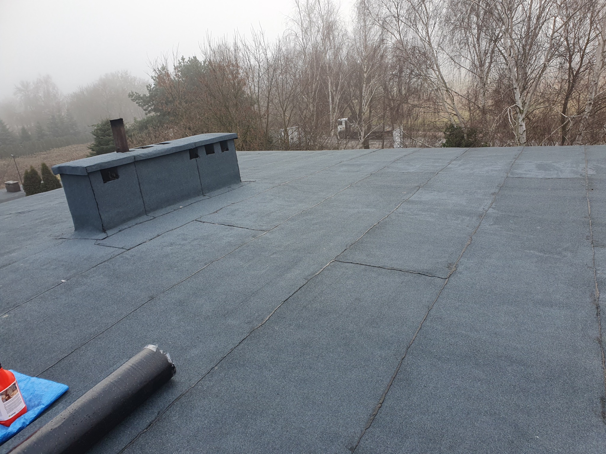 Widok z góry na płaski dach pokryty papą termozgrzewalną, z widocznym kominem i drzewami w tle, z rolką papy i butelką kleju na pierwszym planie.