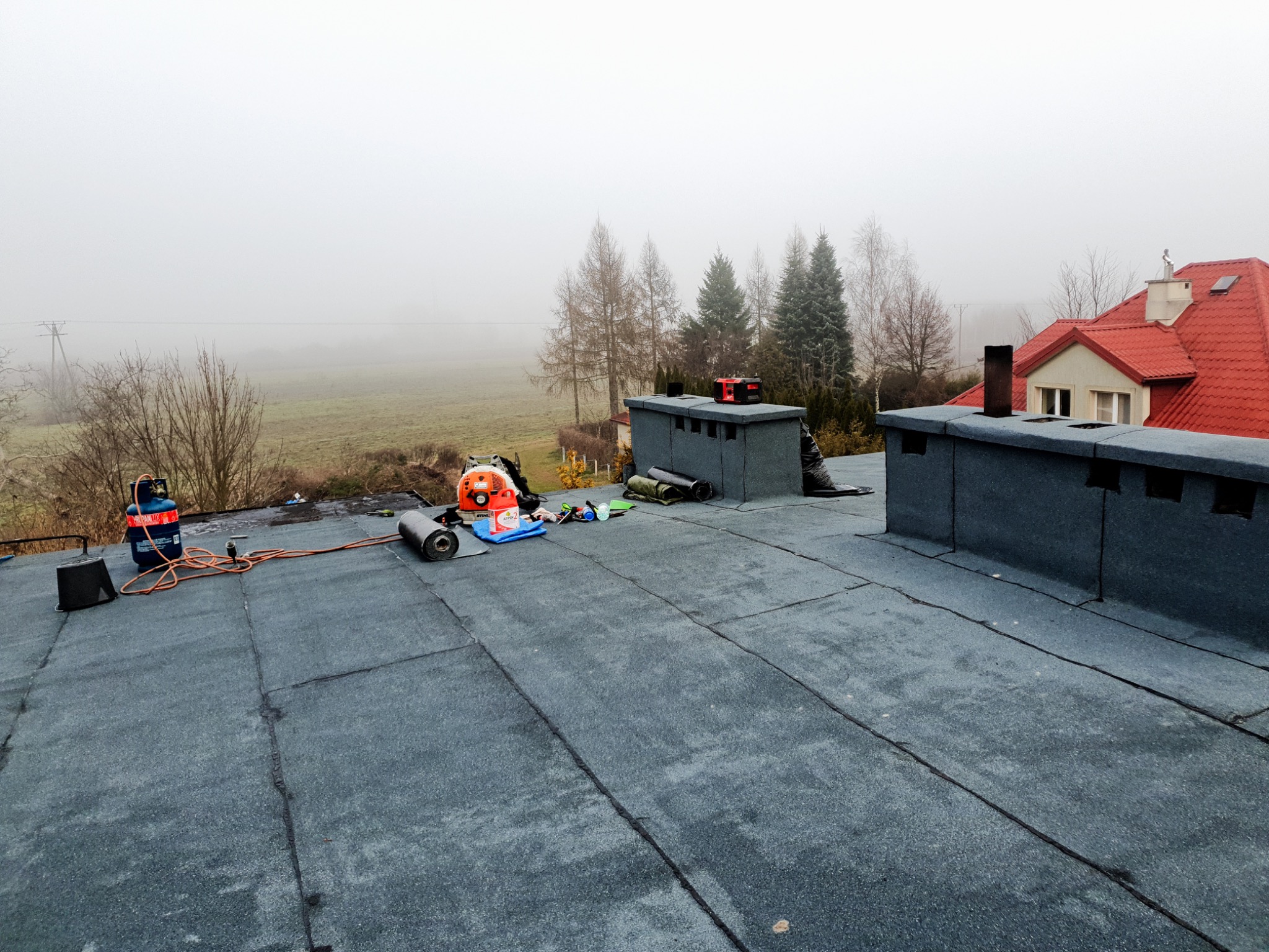 Widok z góry na płaski dach pokryty papą termozgrzewalną, z widocznym sprzętem dekarskim: butlą gazową, rolkami papy, dmuchawą dekarską; w tle zabudowa kominowa i zarys budynku w porannej mgle.
