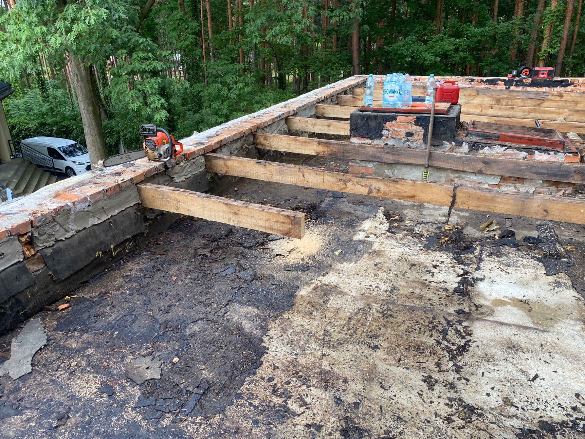 Demontaż starego dachu z widocznymi elementami konstrukcji drewnianej, ceglaną obmurówką, piłą spalinową i butelkami wody mineralnej na tle zielonego lasu.