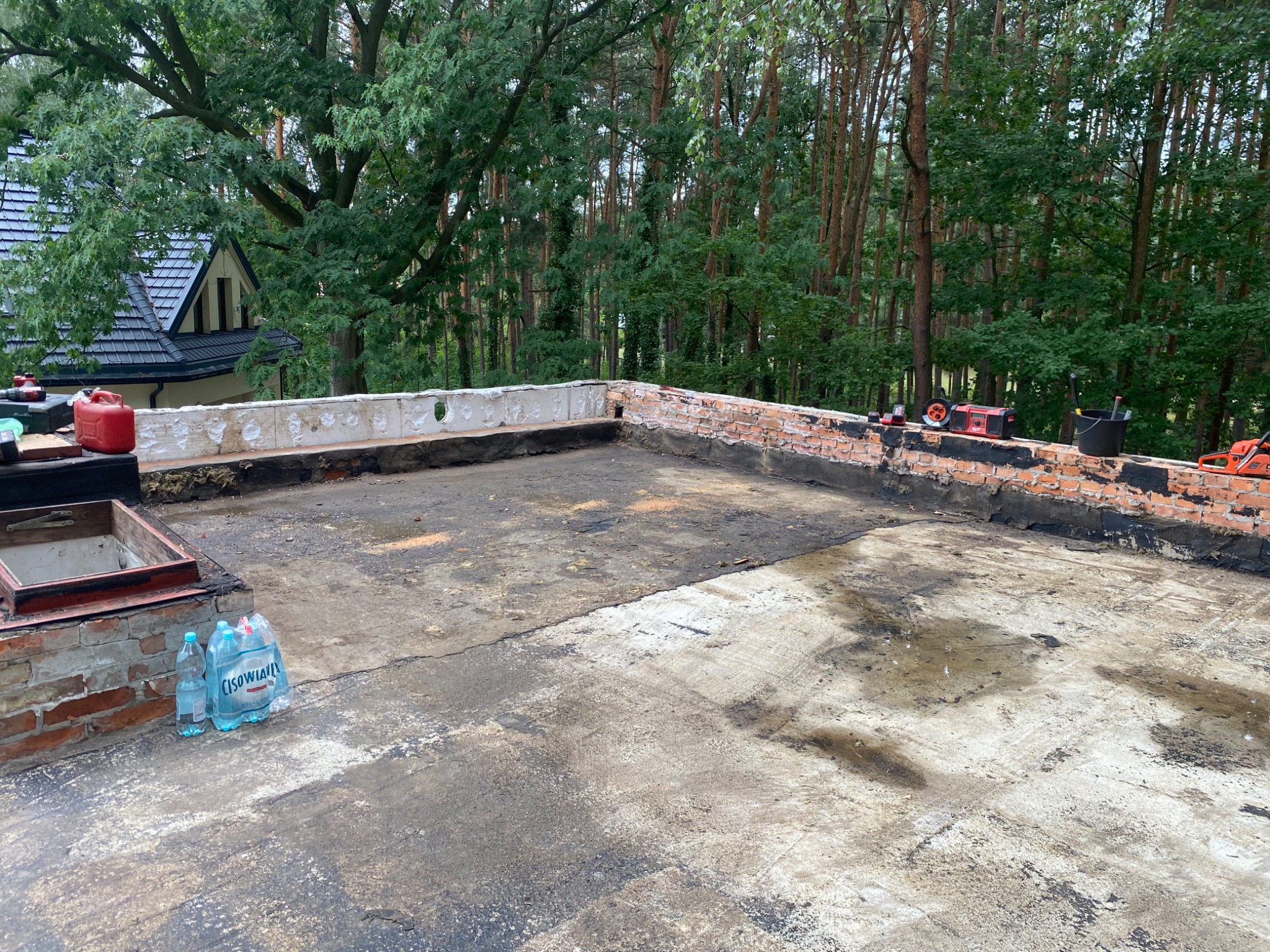 Widok z góry na płaski dach w trakcie prac, z widocznymi narzędziami, butelkami wody mineralnej i lasem w tle; częściowo usunięta stara papa i ceglana attyka.