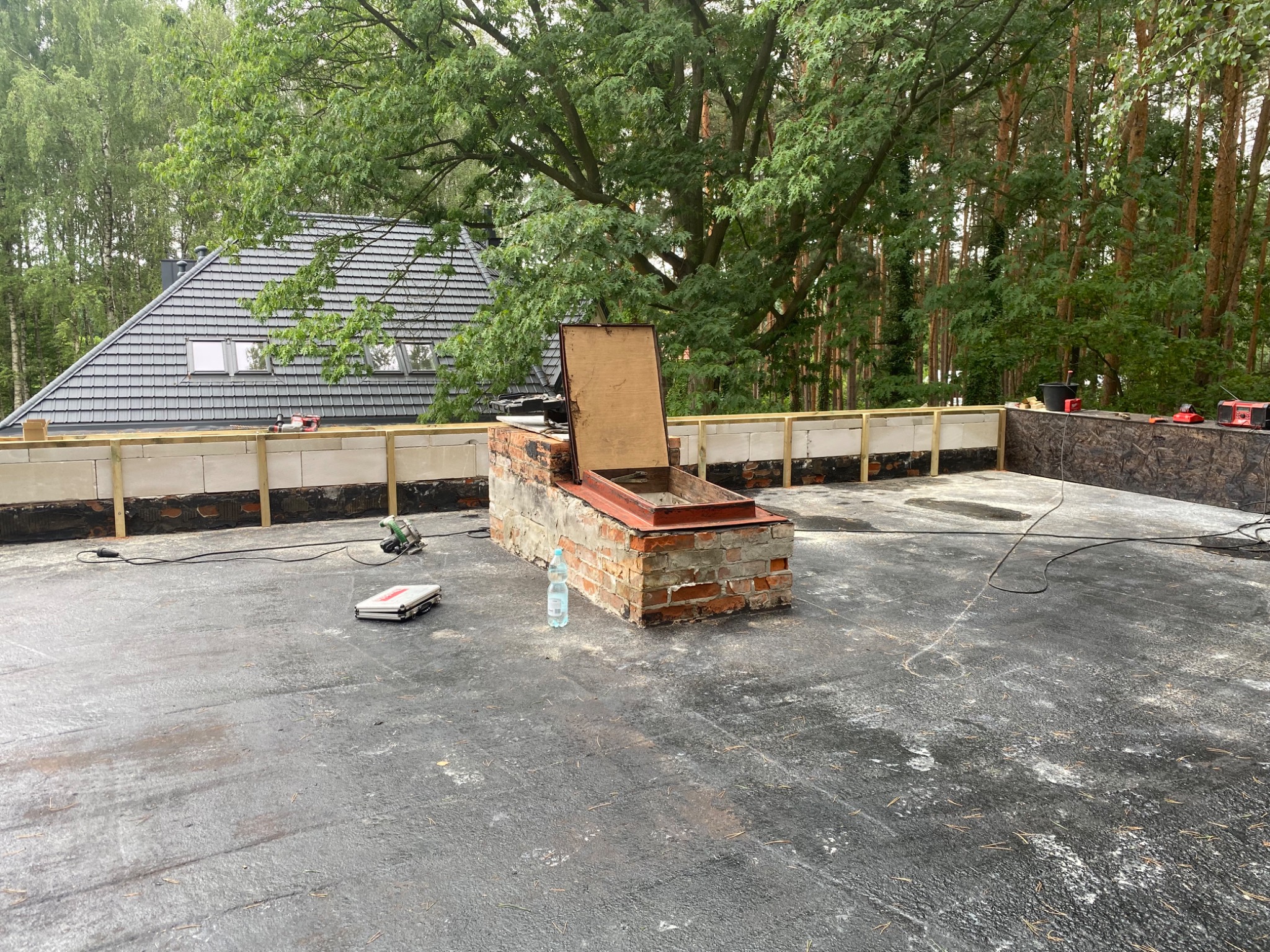 Płaski dach pokryty papą w trakcie budowy, widoczne elementy konstrukcyjne, komin wentylacyjny z otwartym włazem, narzędzia i materiały budowlane, w tle dom z szarym dachem i drzewa.