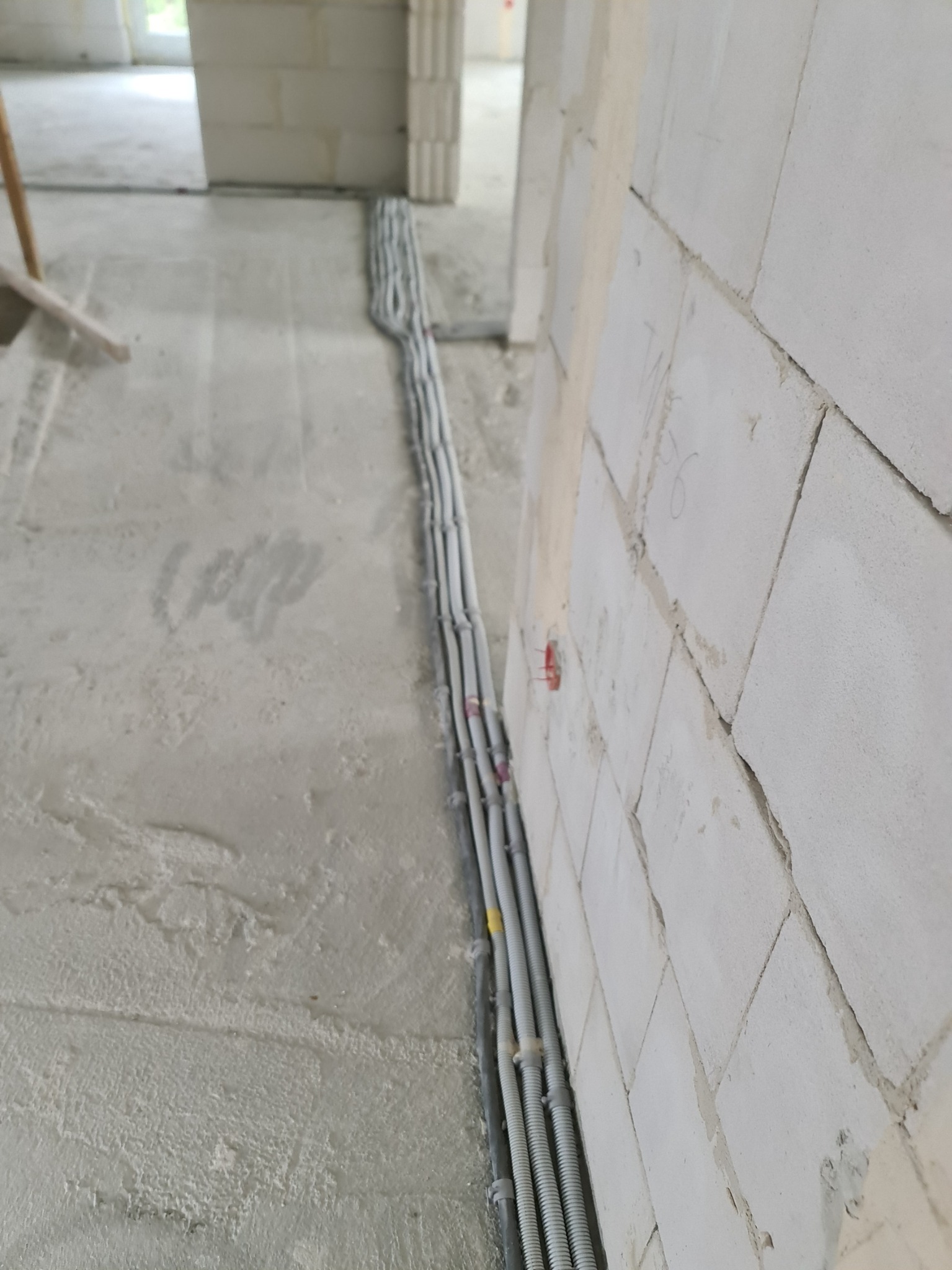 Szereg szarych rurek karbowanych ułożonych wzdłuż ściany z białych bloczków betonowych w surowym wnętrzu budynku w trakcie budowy. Widoczne wystające kable z czerwonymi końcówkami.