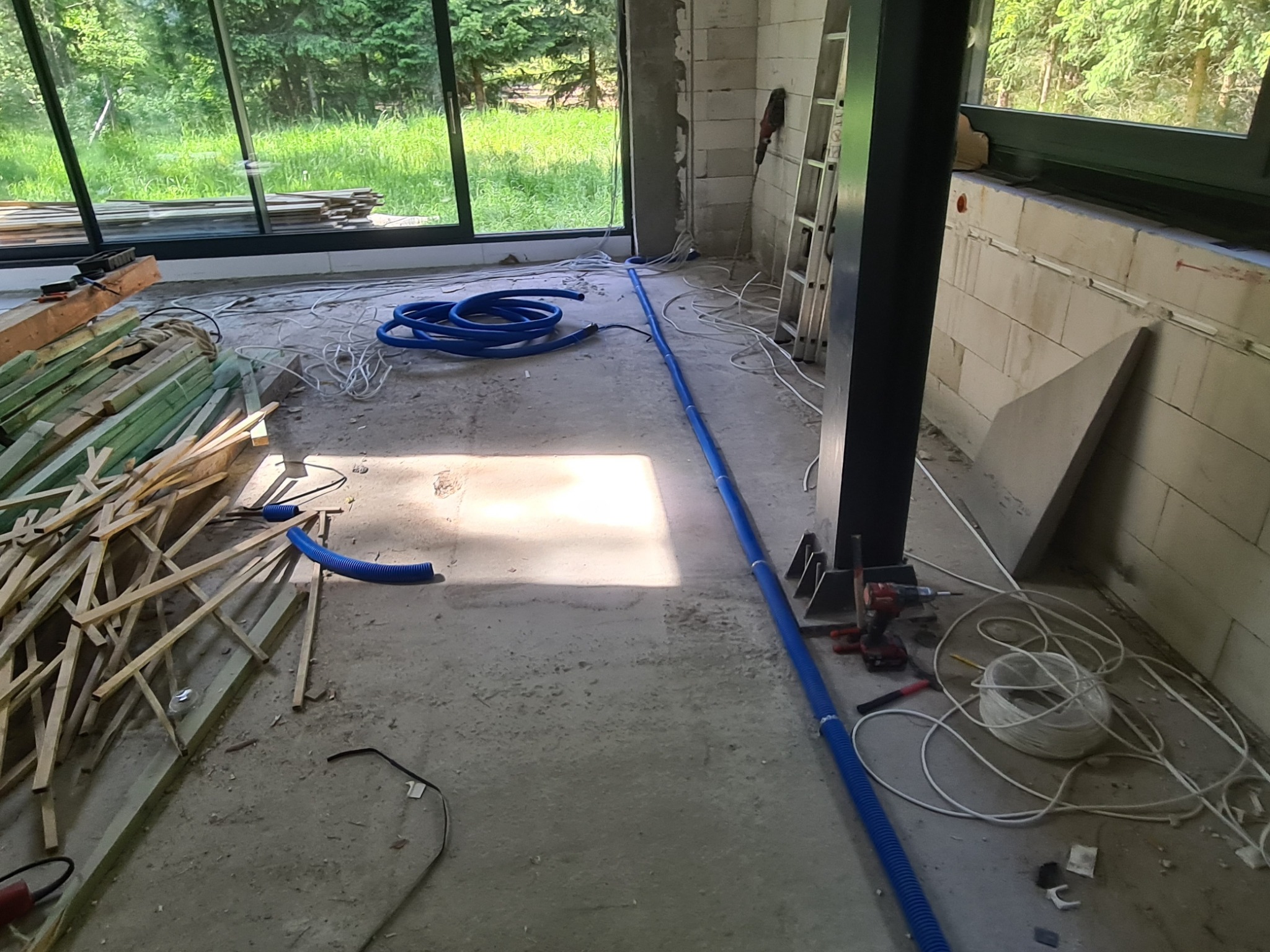 Wnętrze w trakcie prac wykończeniowych: widoczne nieotynkowane ściany z bloczków, okna z widokiem na zieleń, niebieskie rury karbowane, przewody elektryczne, drabina, wkrętarka i drewniane elementy...