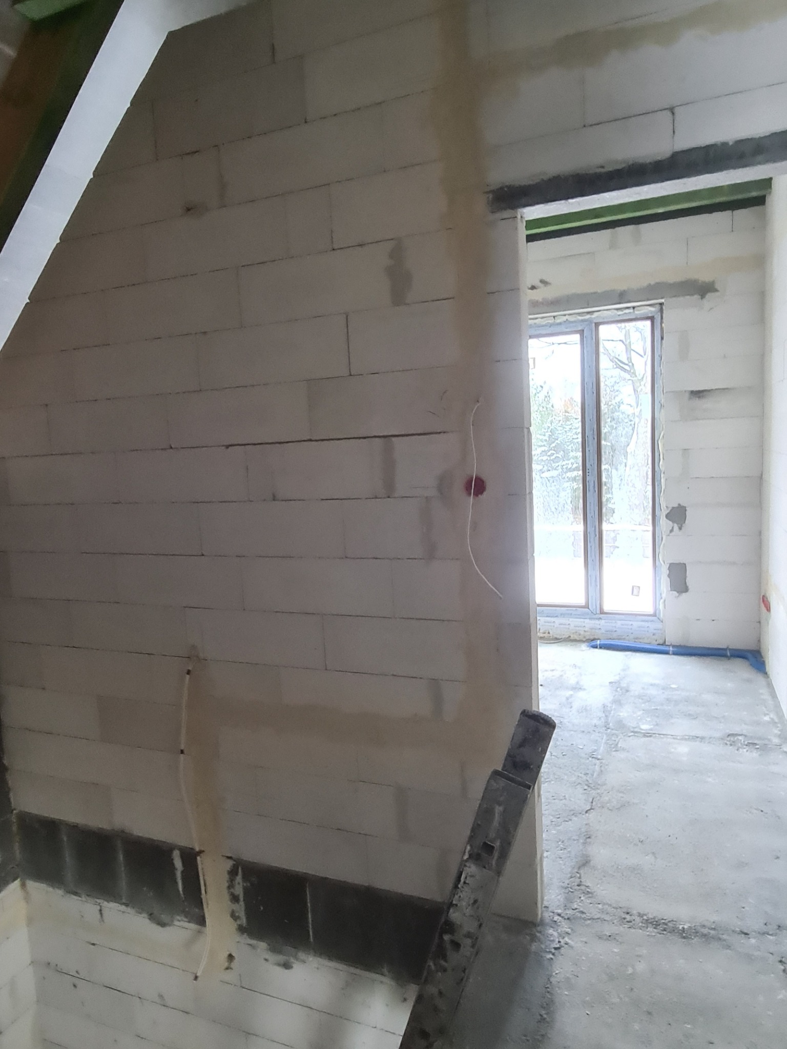 Surowy stan wnętrza w trakcie budowy: ściany z bloczków, widoczne przewody elektryczne wystające ze ściany, okno z widokiem na zewnątrz, betonowa podłoga.