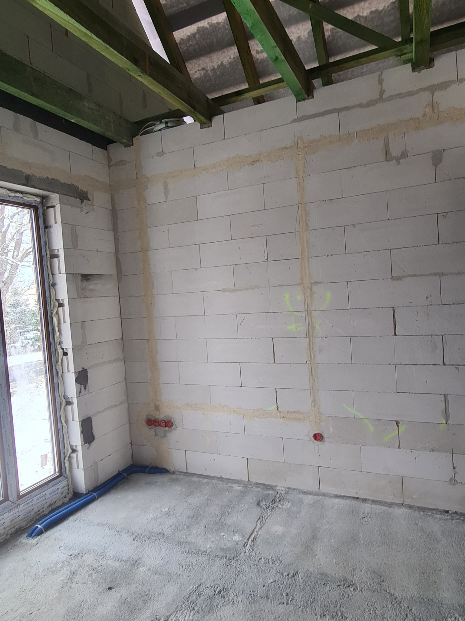 Surowy stan wnętrza z widocznymi bruzdami pod kable elektryczne w ścianie z bloczków, oznaczone trasy, oraz wystające puszki instalacyjne, niebieskie rury karbowane na podłodze.