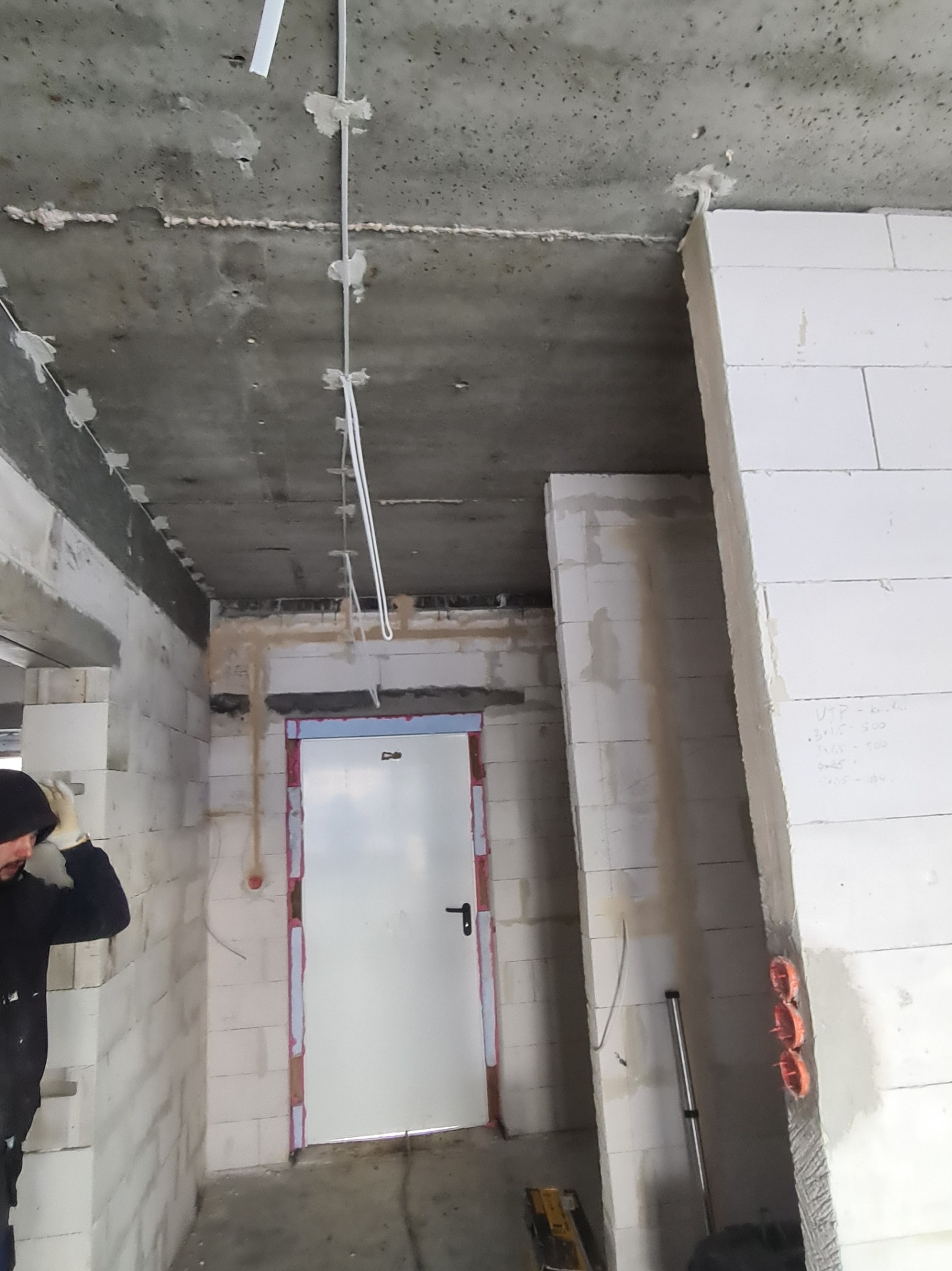 Surowy stan wnętrza w trakcie prac: białe ściany z betonu komórkowego, widoczne przewody elektryczne mocowane do sufitu, puszki elektryczne w ścianie, fragment sylwetki osoby w roboczym ubraniu.