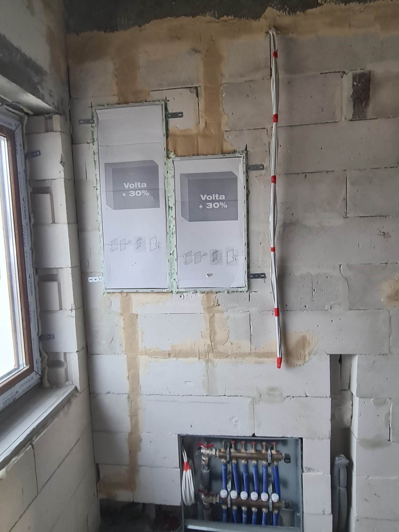 Wnętrze w trakcie instalacji elektrycznej: widoczne dwa panele rozdzielcze Volta +30% zamontowane na ścianie z betonu komórkowego, pionowy kabel elektryczny z czerwonymi oznaczeniami...