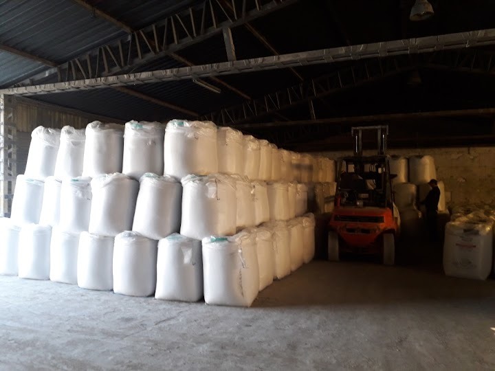 Magazyn z rzędami ułożonych worków z białego tworzywa, w tle wózek widłowy i sylwetka osoby.