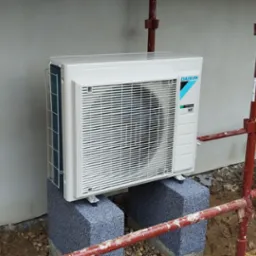 Zewnętrzna jednostka klimatyzacji Daikin zamontowana na dwóch szarych bloczkach betonowych, ustawiona przy ścianie budynku w trakcie prac budowlanych, widoczne rusztowanie i wiadro z kamieniami.