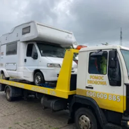 Laweta z ż&oacute;łtym wysięgnikiem przewozi biały samoch&oacute;d kempingowy marki Fendt w pochmurny dzień.