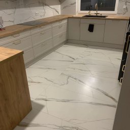 Tomasz Zegzuła - Wykończona kuchnia z białymi szafkami, drewnianym blatem i podłogą z płytek imitujących marmur, widok na okno z żaluzjami.