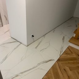 Tomasz Zegzuła - Połączenie dwóch rodzajów podłóg: białe płytki imitujące marmur z wyraźnym użyleniem oraz parkiet ułożony we wzór jodełki francuskiej, widoczny fragment schodów.